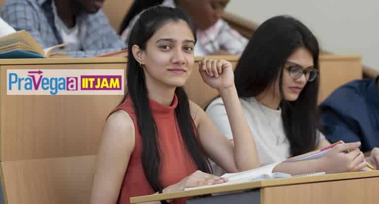 Two female students sit at a classroom desk with notebooks, one glancing at the camera; a 'PraVega ITJAM' sticker is on the desk edge.